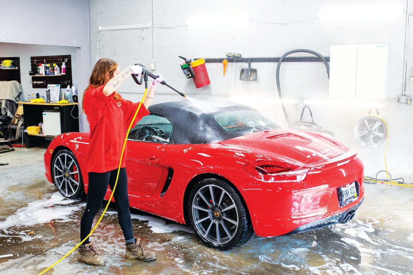 A person in a red shirt uses a pressure washer to clean the black convertible top of a bright red Porsche inside a garage.