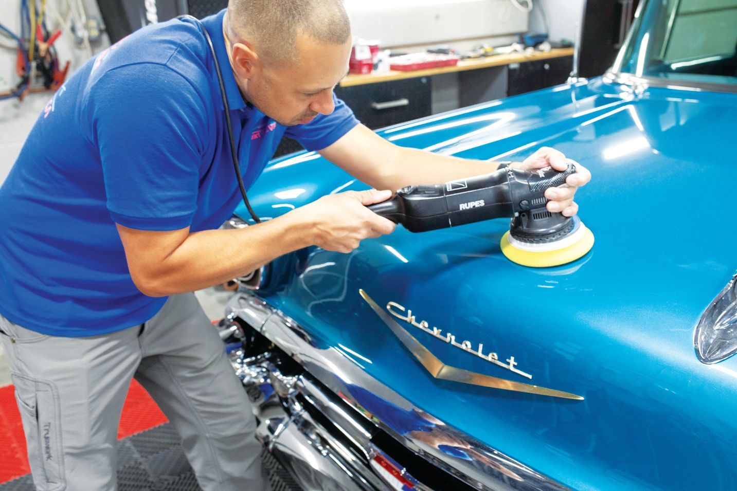 A professional uses a rotary polisher to buff the teal hood of a classic Chevrolet in a workshop.