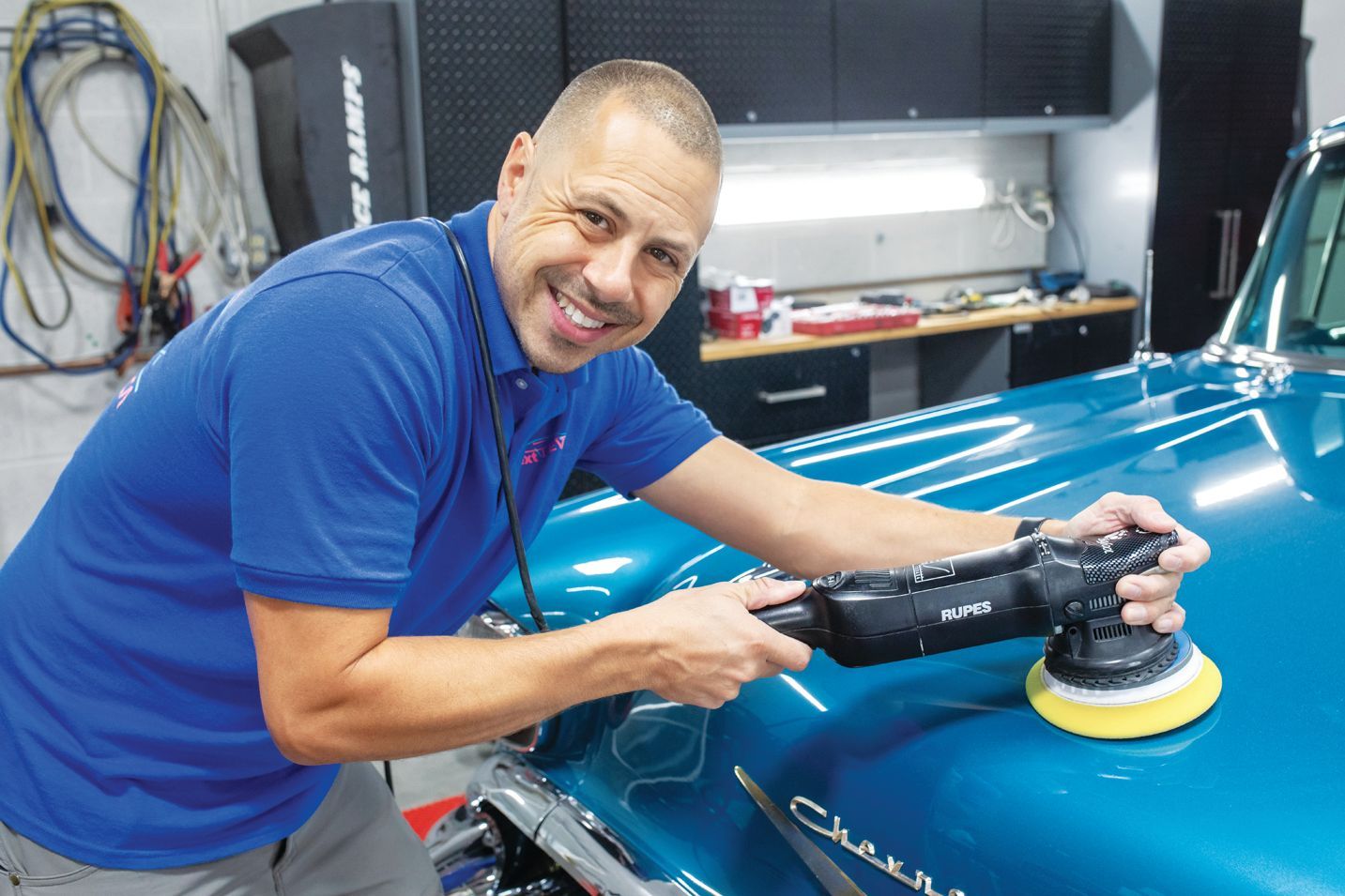 A technician in a blue polo shirt smiles while using a power buffer to polish the teal hood of a classic car in a workshop.