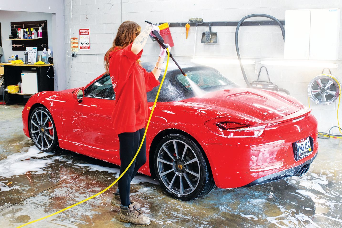 A person wearing a red sweatshirt uses a pressure washer to clean a red sports car in a garage.