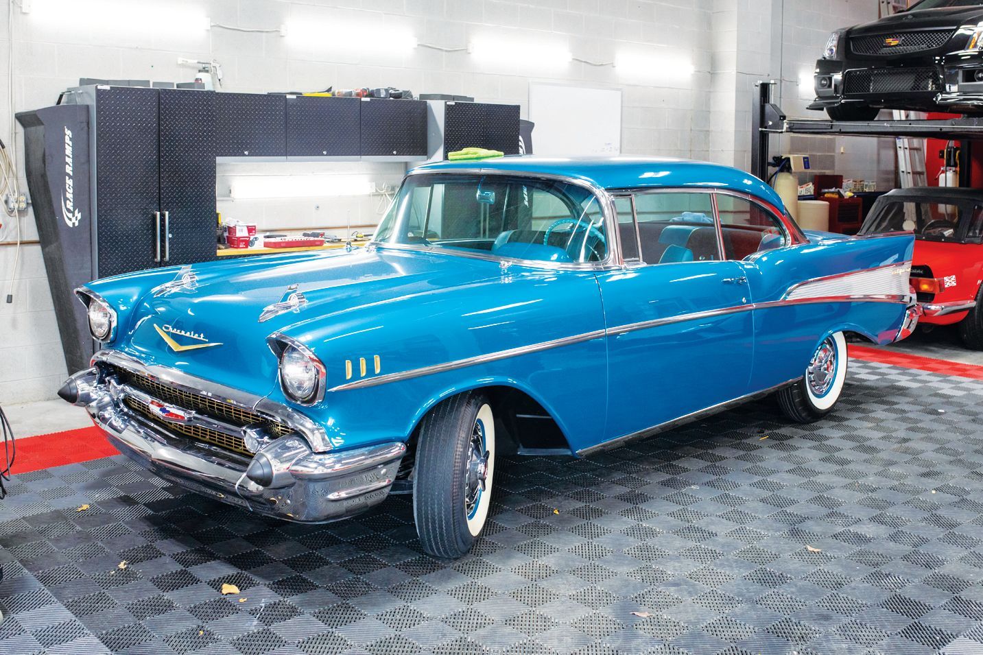 A bright blue vintage Chevrolet Bel Air parked inside a well-lit auto garage with black flooring.
