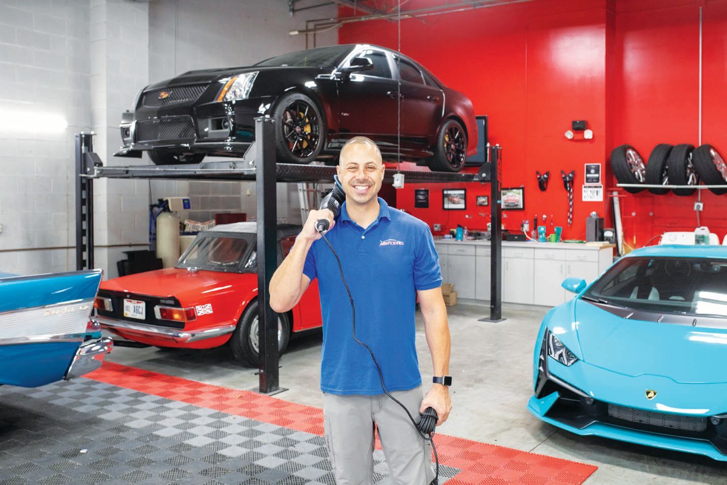A person stands in a car garage with a buffer tool, surrounded by several luxury cars, including a blue Lamborghini.