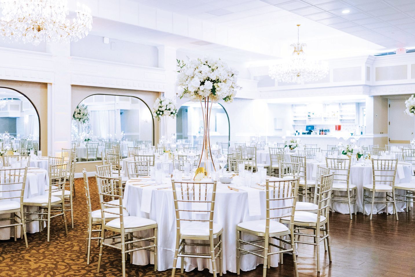 A bright, elegant banquet hall set for a formal event with round tables, white linens, and tall floral centerpieces.