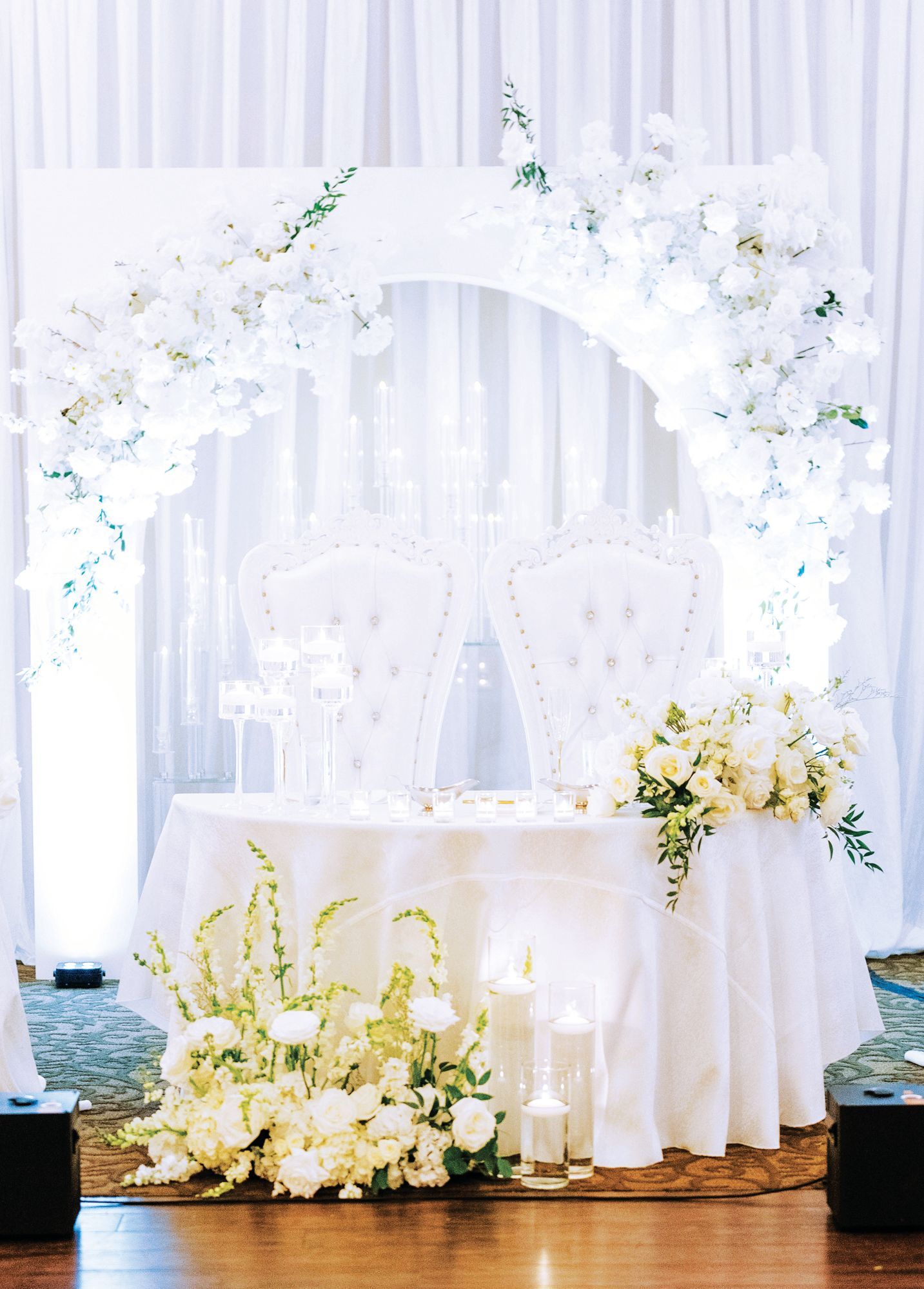 A white wedding sweetheart table with two tufted chairs, arched floral backdrop, and floor candles.