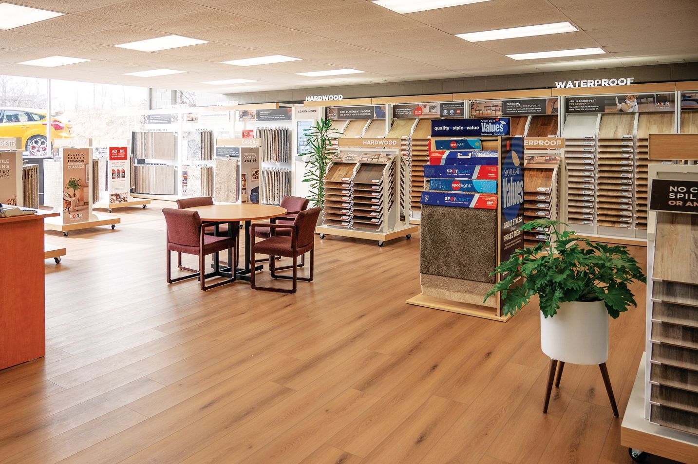 A brightly lit flooring showroom featuring wood-look vinyl planks, display racks with carpet samples, and a seating area.