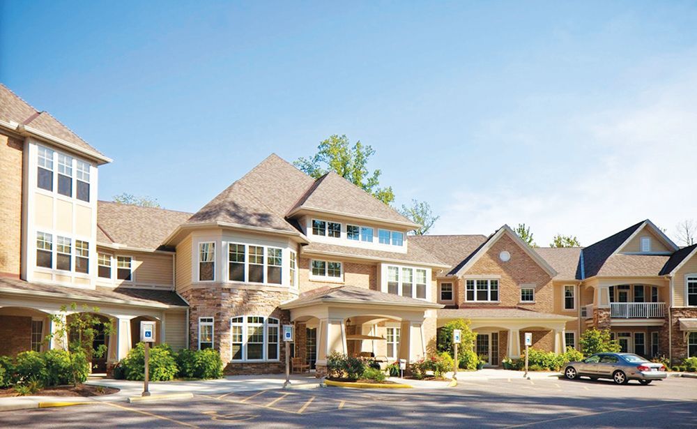 A large, multi-story beige brick building with a pitched roof, featuring a covered entrance and an outdoor parking area.