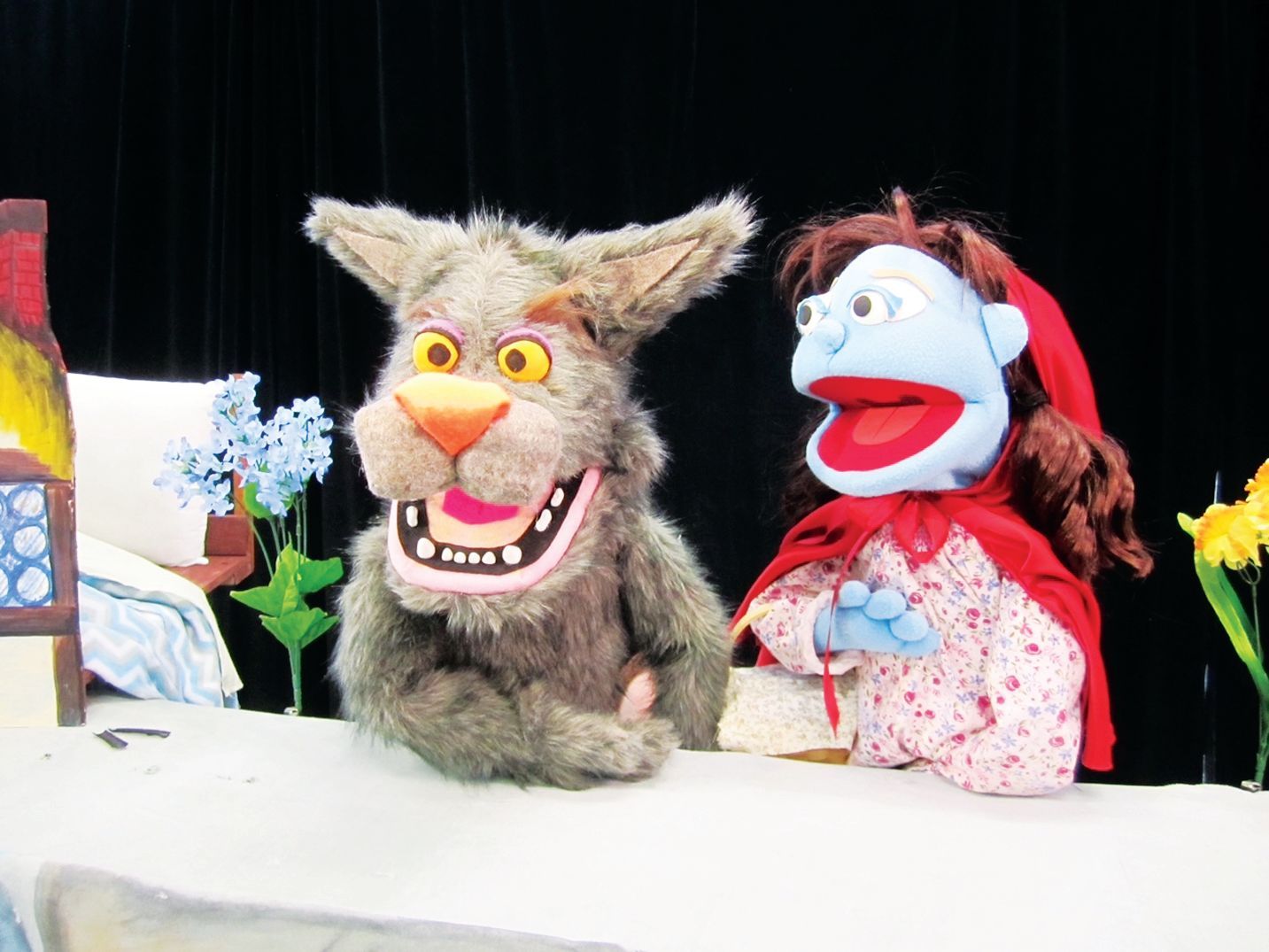Puppet theater scene featuring a gray wolf puppet and a blue-faced human puppet wearing a red hood against a black backdrop.
