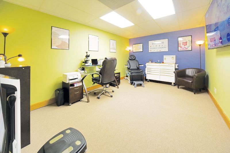 A treatment room with a desk, office chair, exam chair, medical cabinet, and armchair against green and blue walls.