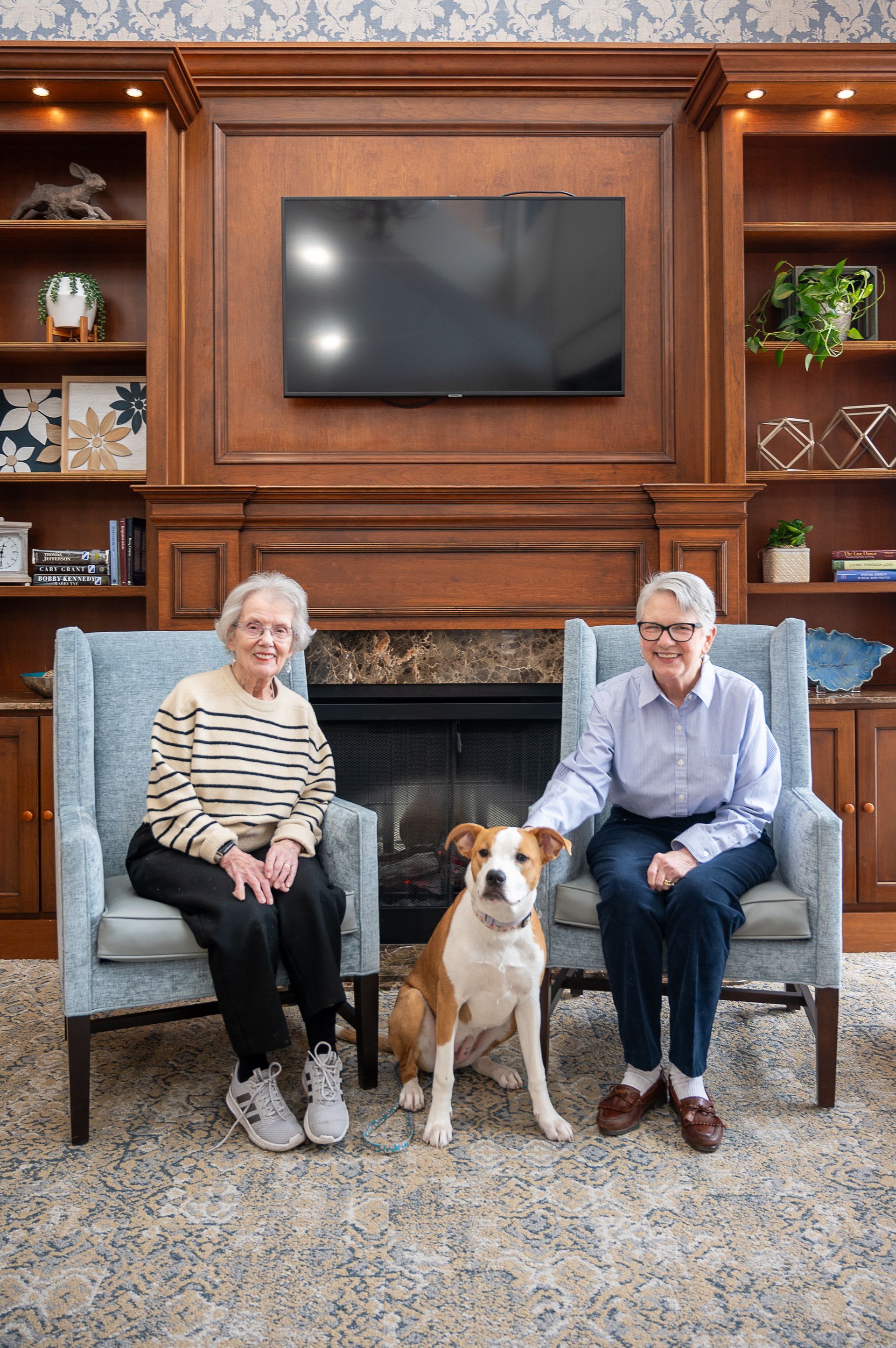 Two people sit in armchairs in front of a fireplace and built-in bookshelves with a dog standing between them.