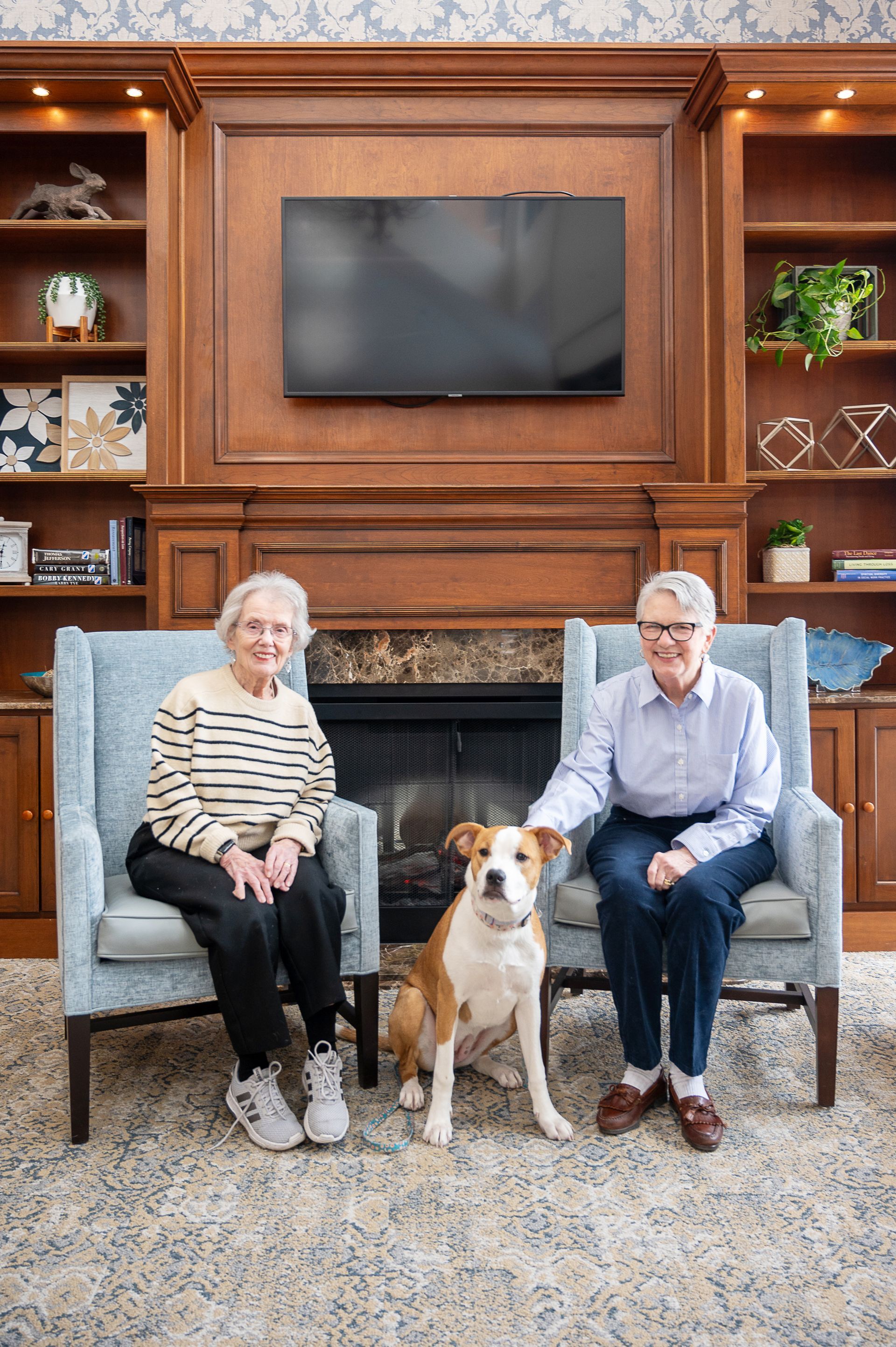 Two people sit in upholstered chairs flanking a fireplace and a large dog, with built-in wooden bookshelves behind them.