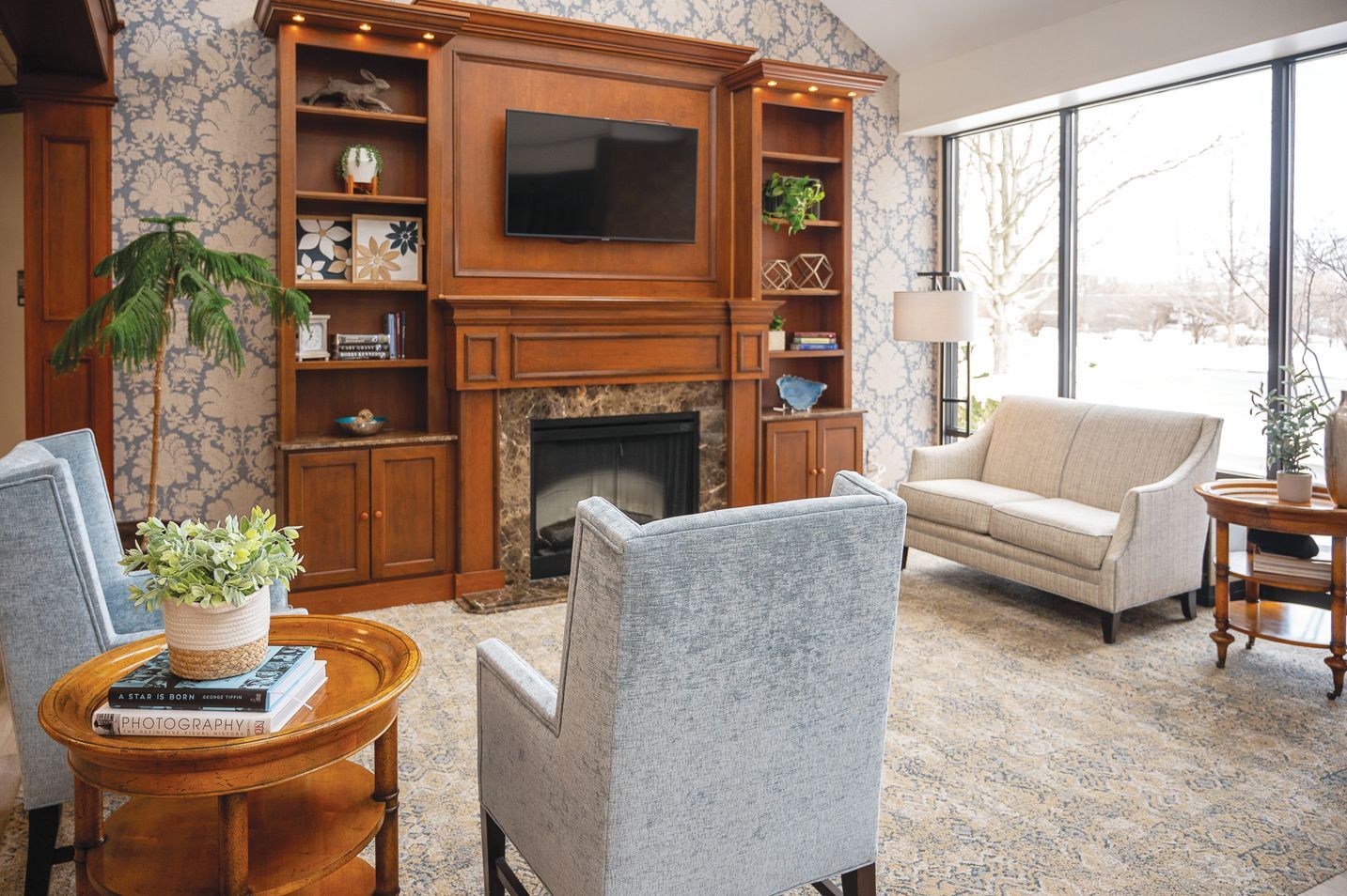 A cozy living room with patterned wallpaper, a central wooden fireplace, two blue armchairs, and a beige sofa.