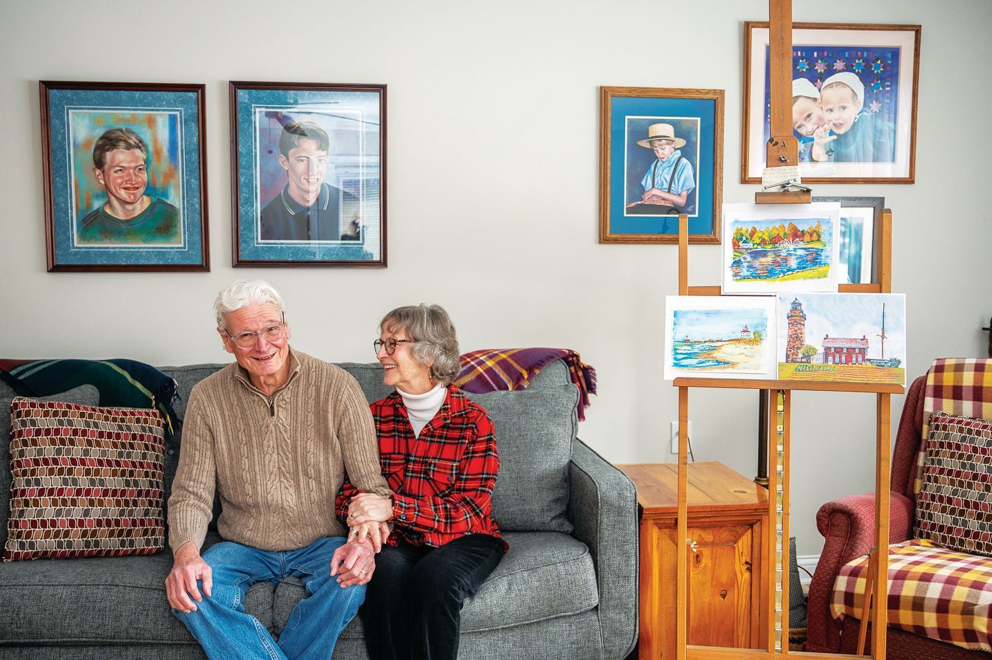 A couple sits on a grey couch in a room decorated with various paintings on the wall and an easel displaying artwork.