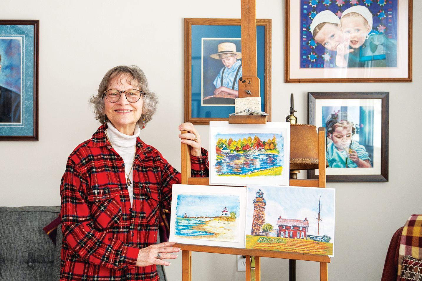 A person smiling in a red plaid shirt stands next to an easel displaying three of their own watercolor paintings.