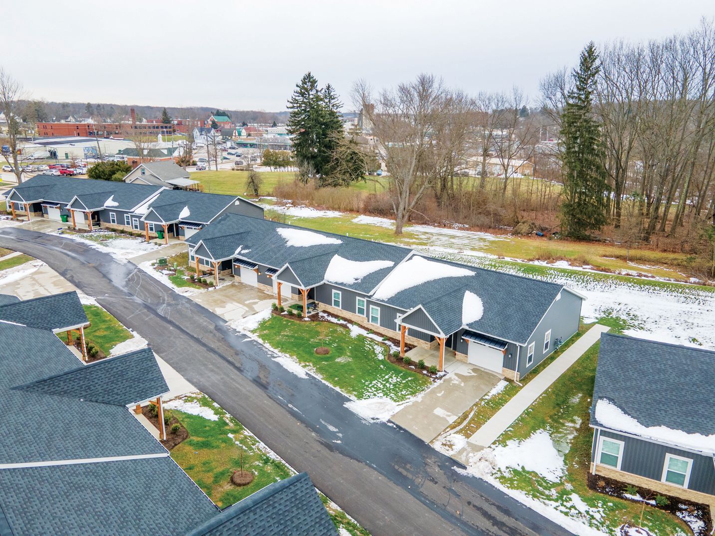 Aerial view of a residential complex with dark-roofed buildings, a central asphalt road, and light snow on the grounds.