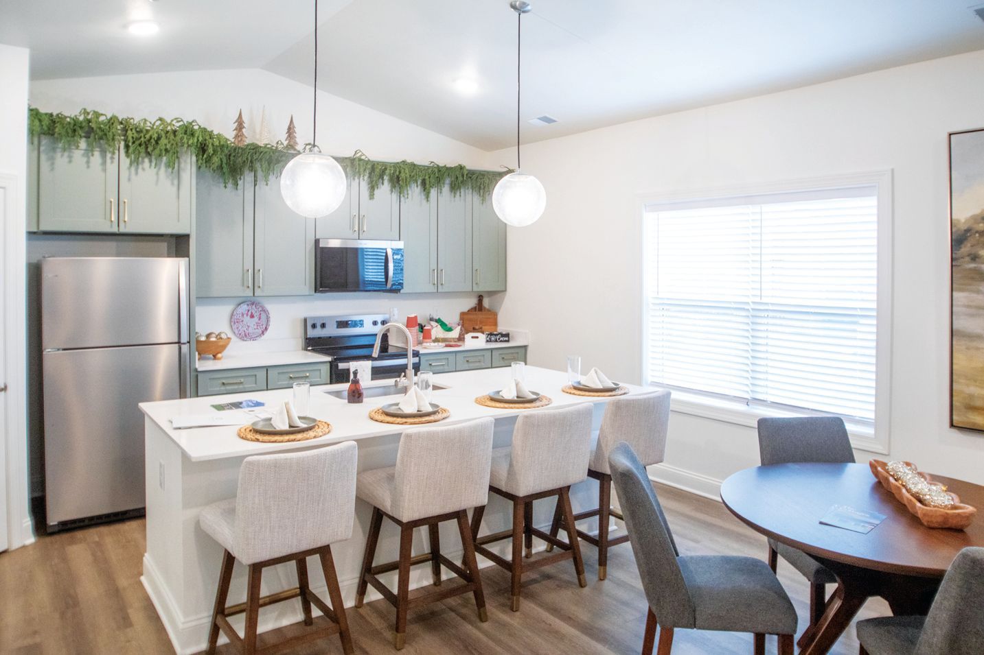 A modern kitchen with sage green cabinets, stainless steel appliances, a white island with bar stools, and a dining table.