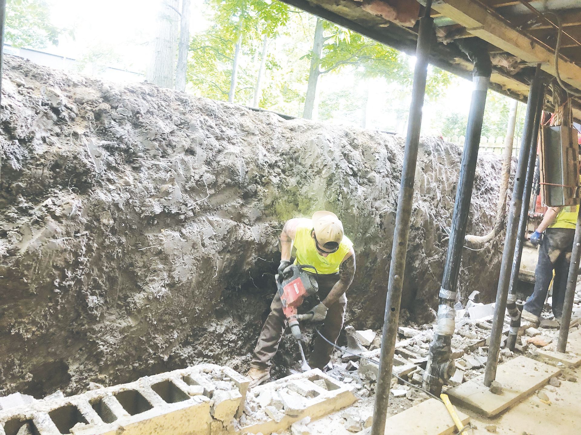 A construction worker in a high-visibility vest uses a jackhammer to break up concrete next to a deep soil trench.