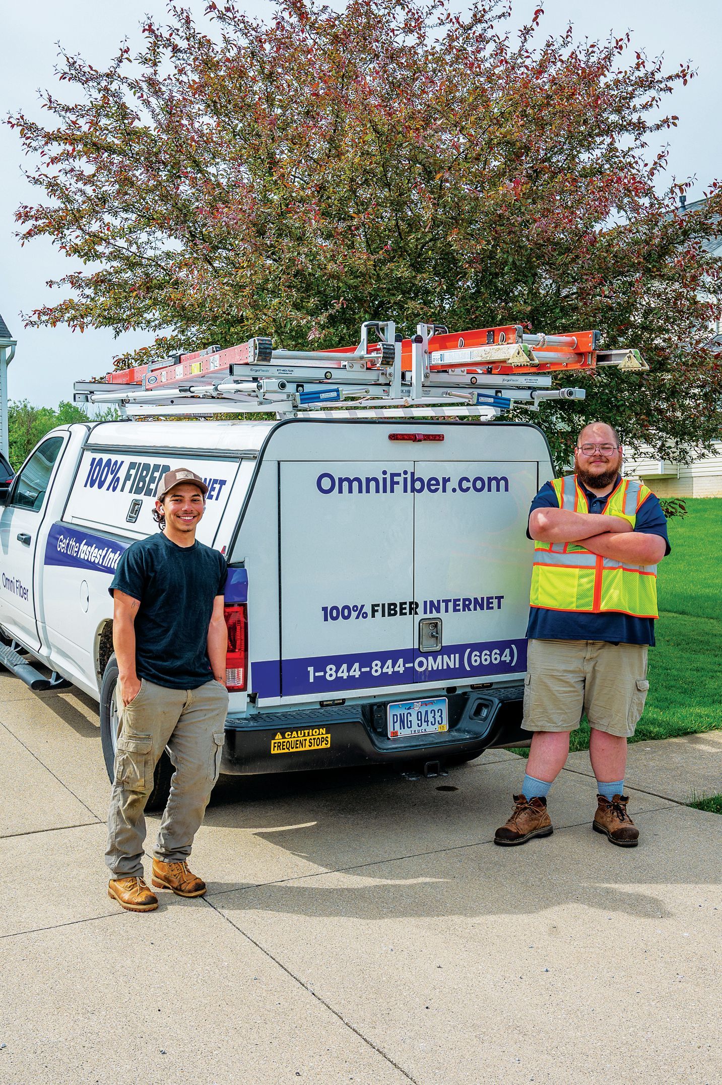 Two technicians stand in front of an OmniFiber work truck with ladders on the roof, positioned before a leafy tree.
