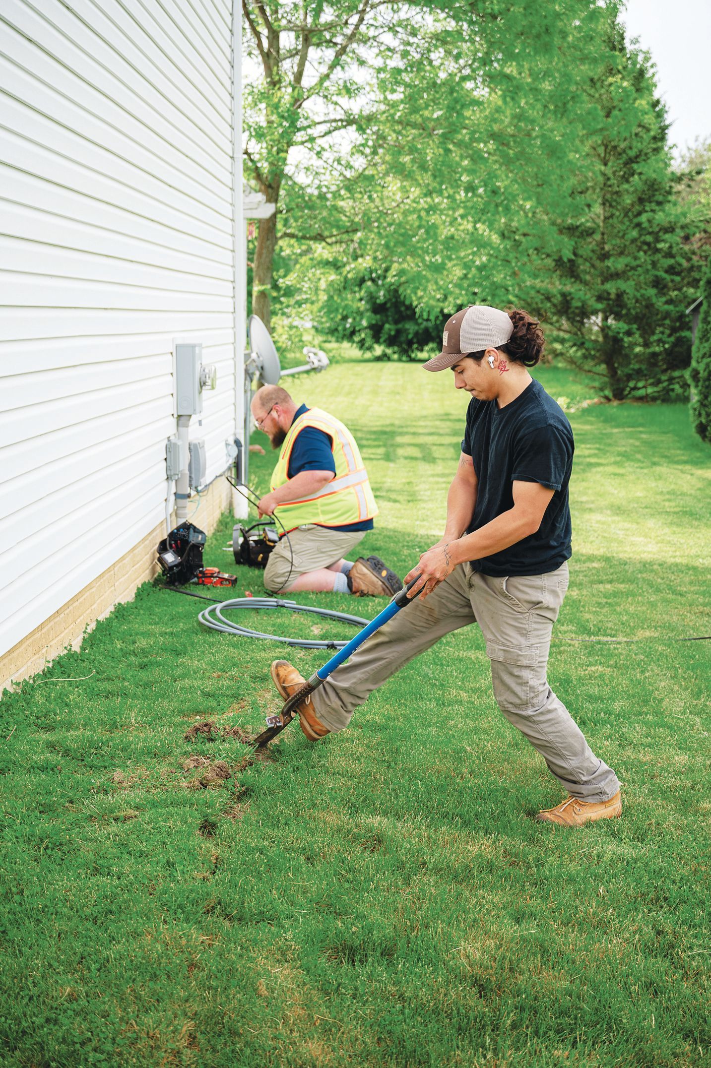 Two workers install outdoor equipment near the side of a building; one uses a shovel while the other kneels nearby.
