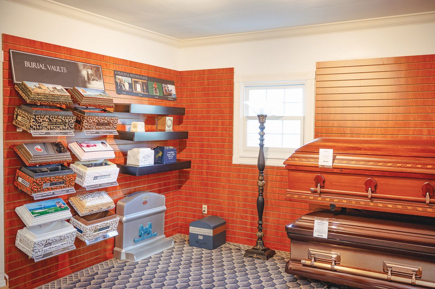 A display room with wooden caskets, shelves of funeral urns, and a memorial monument against orange brick-patterned walls.