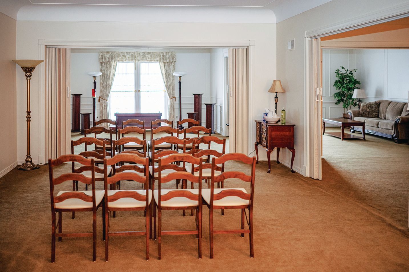 Rows of wooden chairs face a brightly lit, curtained doorway in a beige-carpeted meeting room, with a lounge area nearby.
