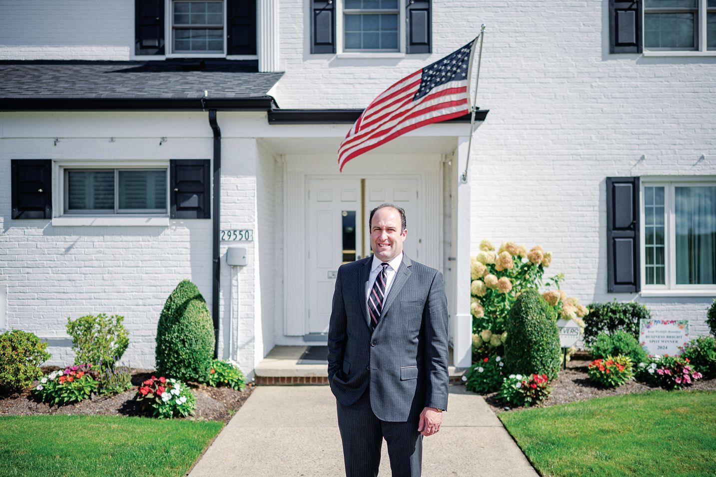 A person in a suit stands on a walkway in front of a white brick building with an American flag above the entrance.