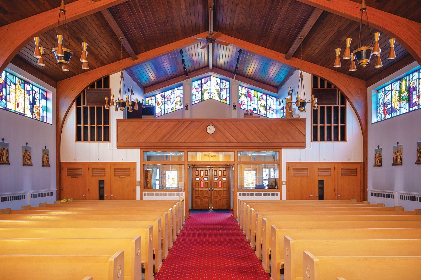 The interior of a church with rows of wooden pews, a red carpeted aisle, stained-glass windows, and vaulted wood ceilings.