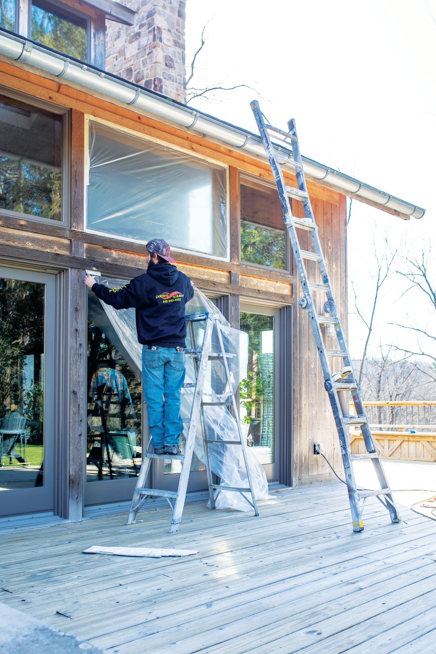 A person on a step ladder working on the exterior trim of a wooden home, with a tall extension ladder leaning nearby.