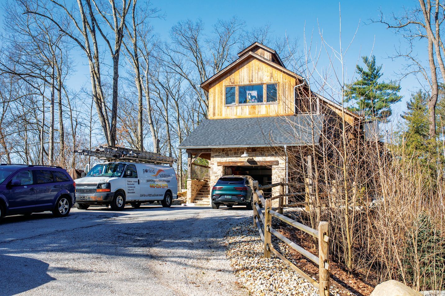 A work van and two cars parked on a gravel driveway in front of a wood and stone house on a sunny day.