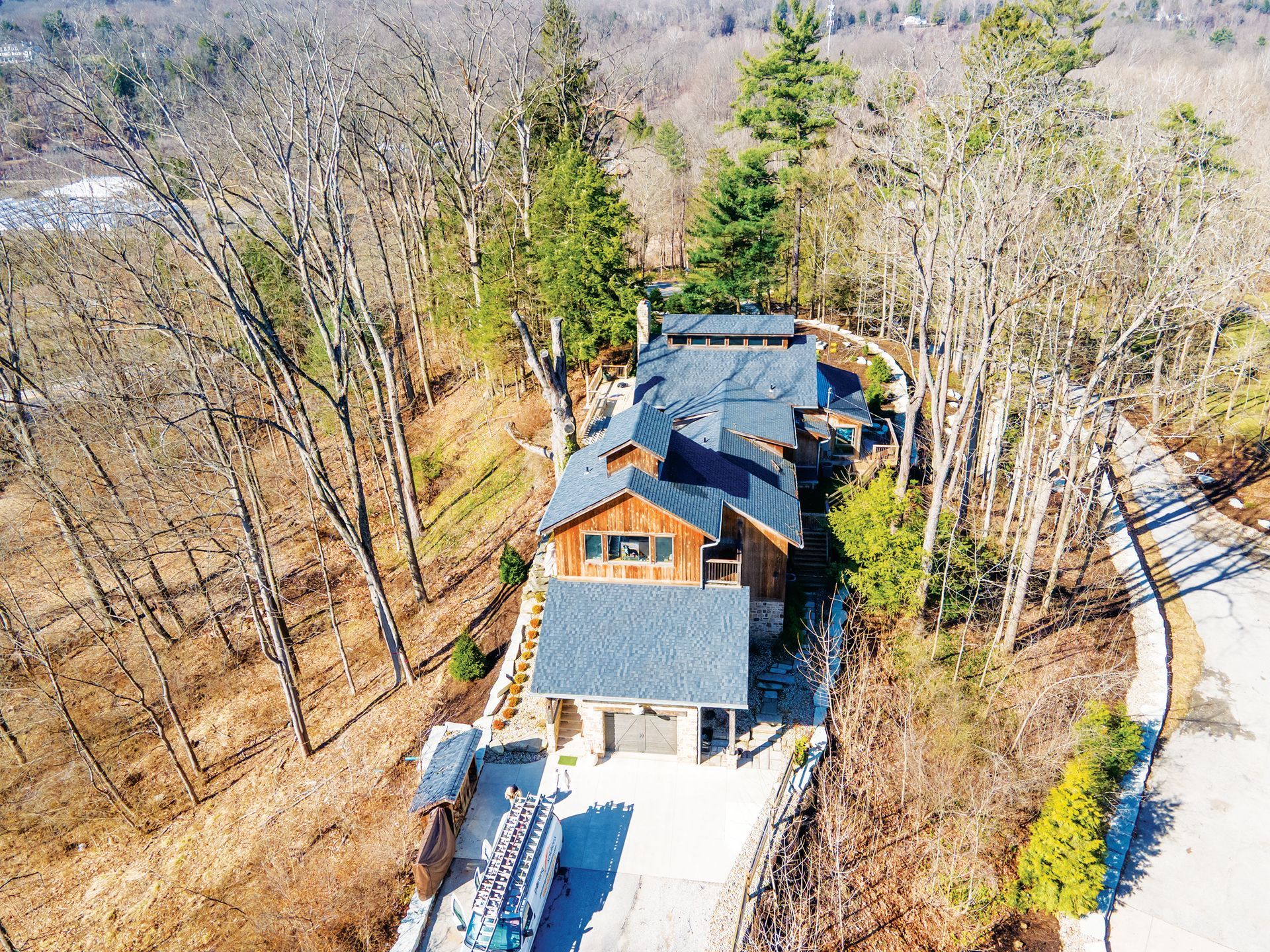 A multi-level wood and stone home nestled among bare trees on a sunny hillside with a paved driveway.