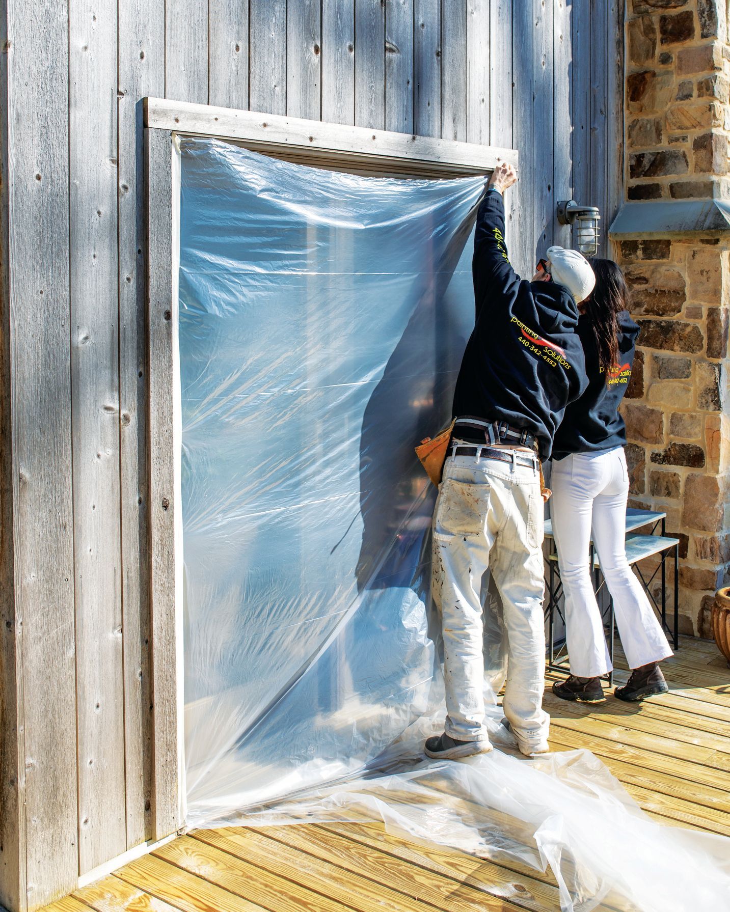 Two people in work clothing attach a large, clear plastic sheet over a doorway on a rustic wooden building exterior.