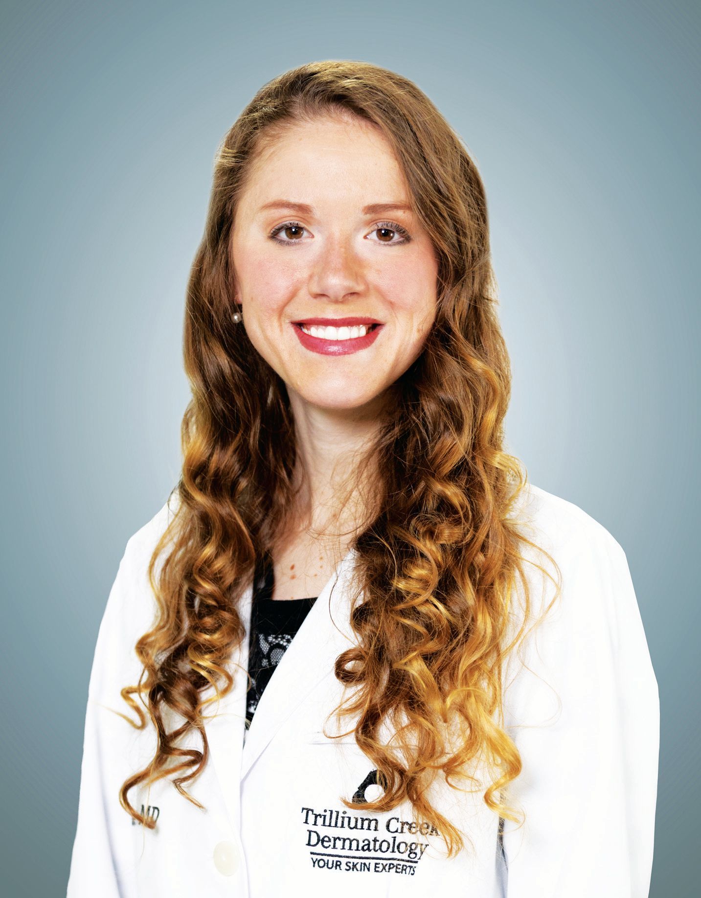 A professional headshot of a person in a white lab coat with a Trillium Creek Dermatology logo on a blue background.