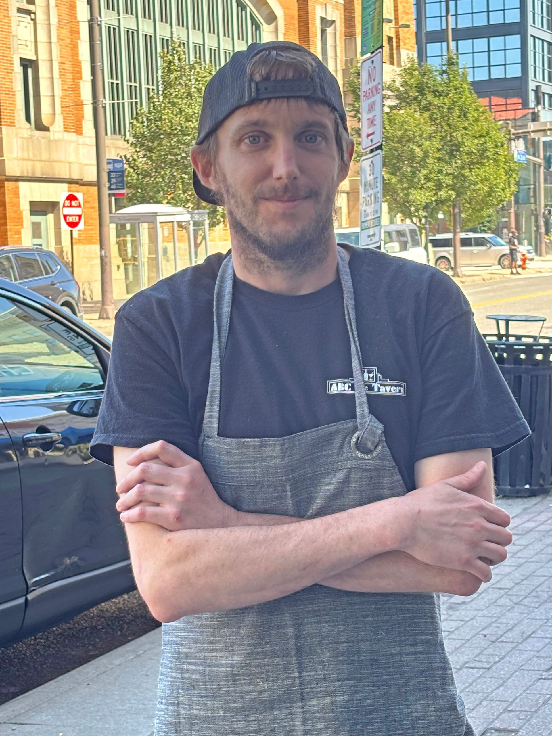 A person with a beard and baseball cap, wearing a dark shirt and apron, stands with arms crossed on a sunny city street.