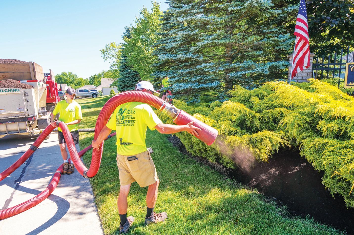 Two workers in neon yellow shirts use a large red hose to spread mulch onto a garden bed beside a house with a US flag.