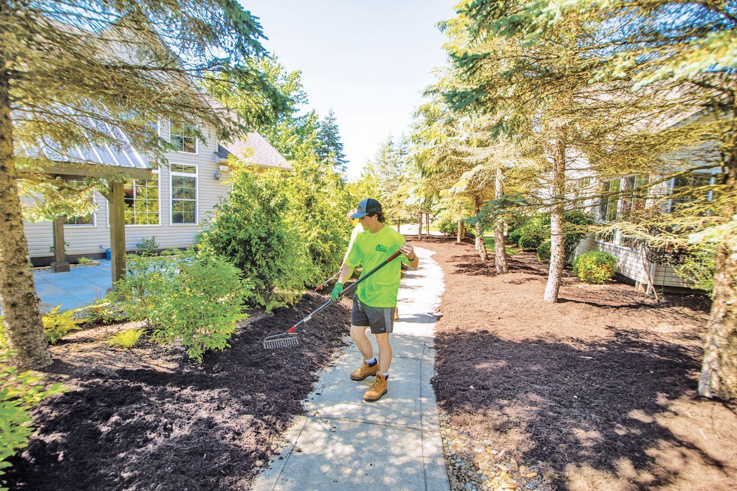 A landscaper in a neon green shirt spreads mulch along a stone garden path between two houses.