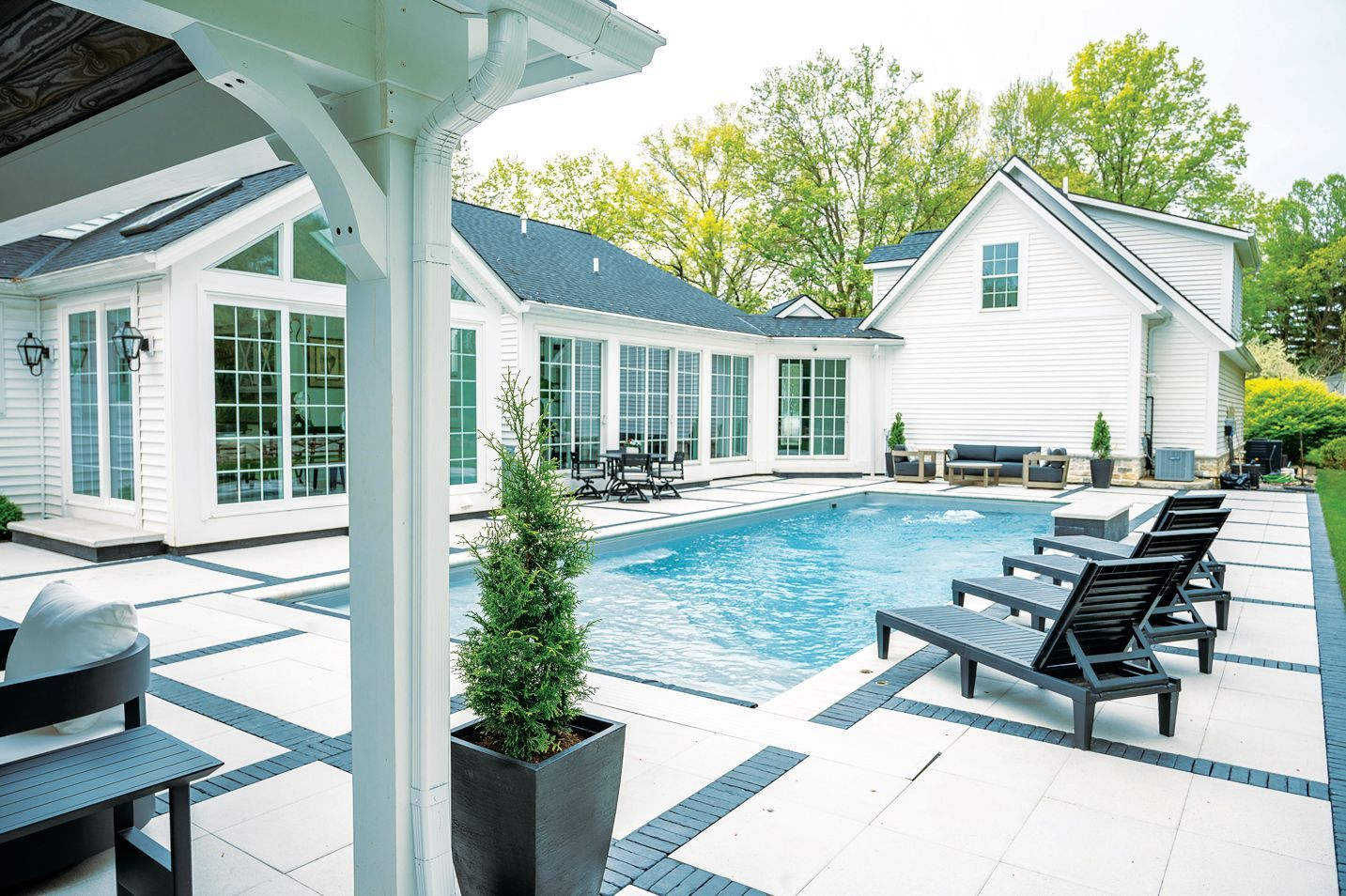 A white house with a blue-roofed patio, swimming pool, and lounge chairs on a paved deck.