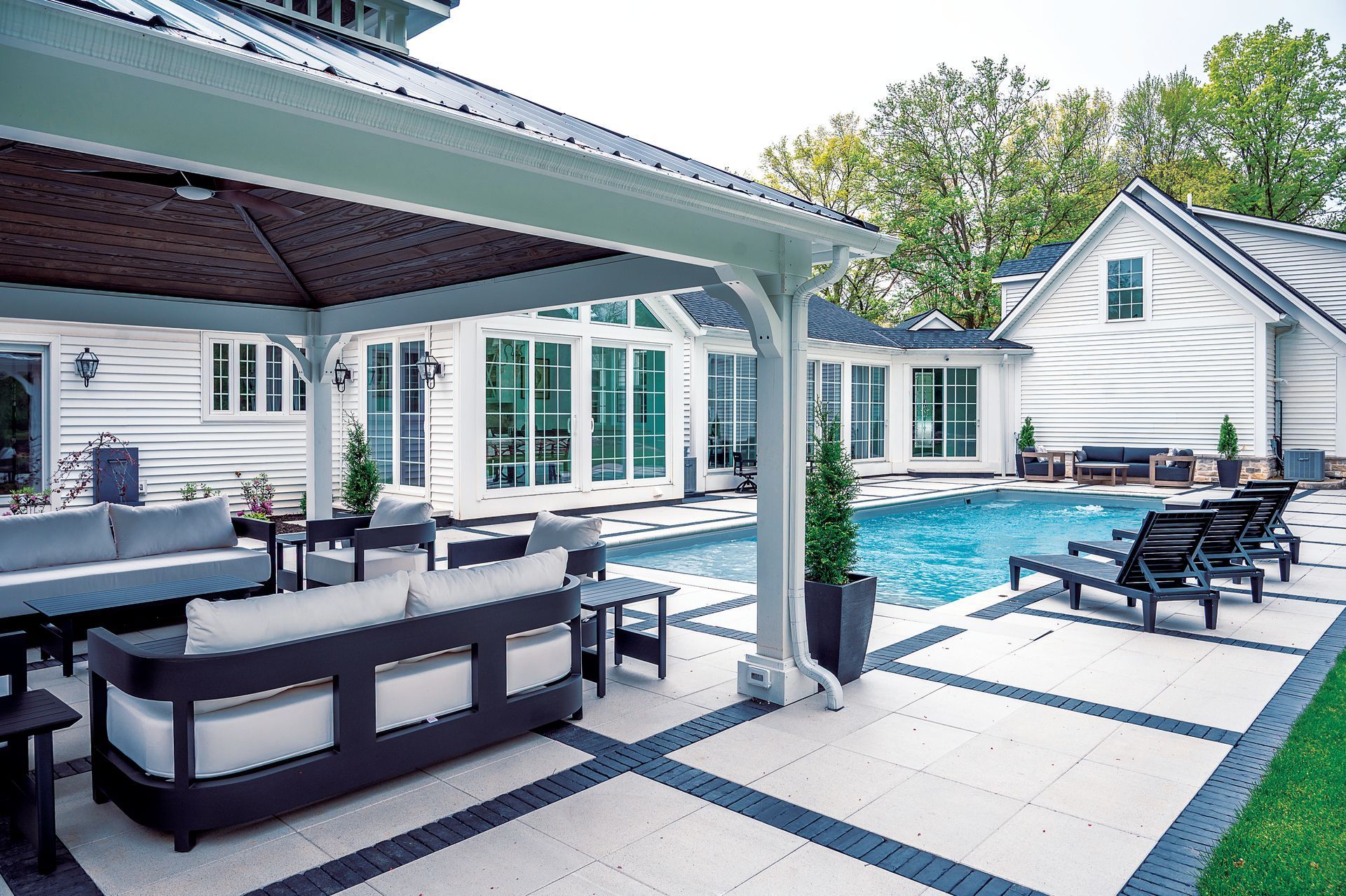 Outdoor patio with a shaded seating area, lounge chairs, and a swimming pool next to a white house with dark trim.