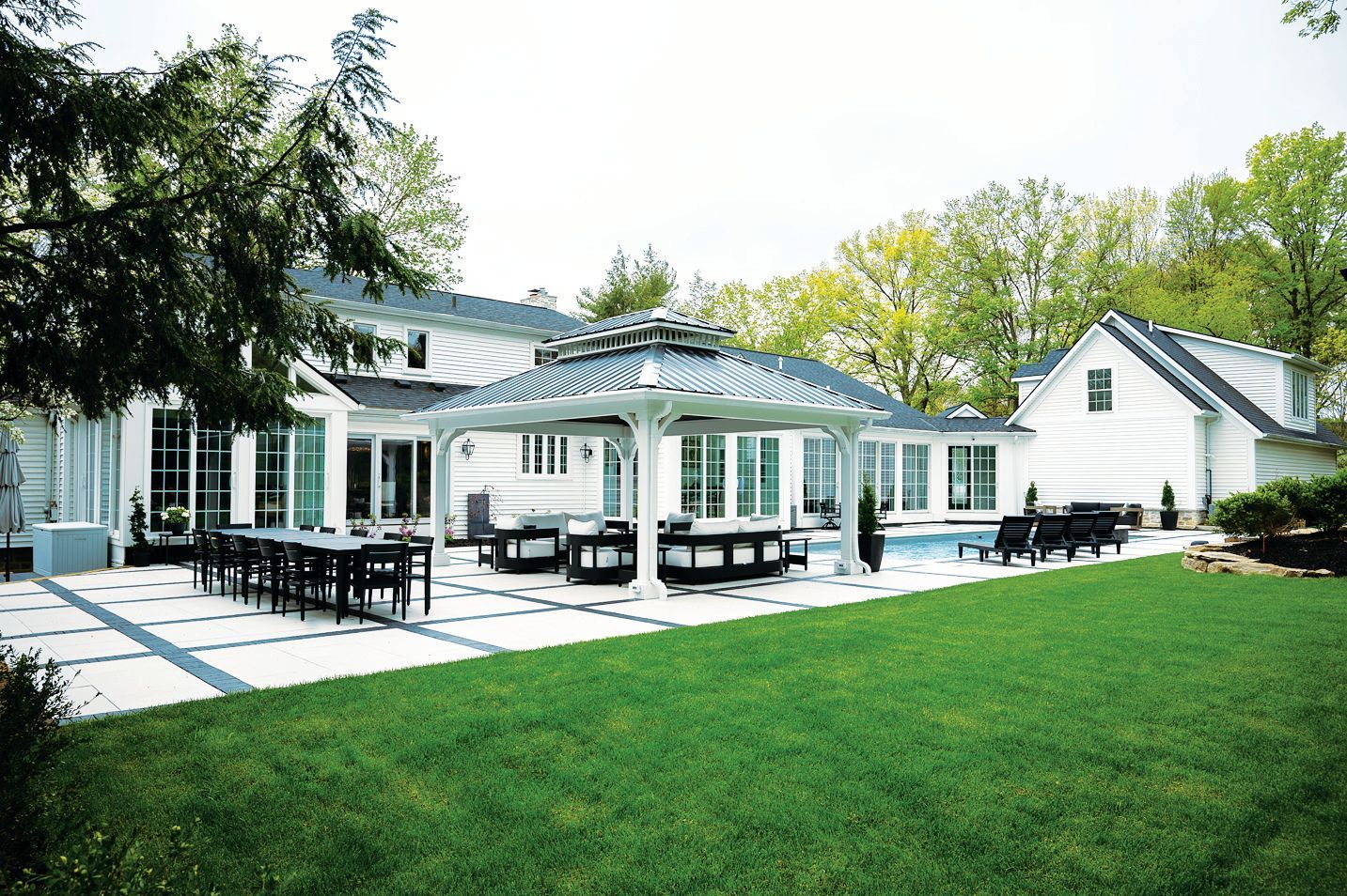 White house with a gray-roofed gazebo, pool, and patio dining set in a grassy backyard with trees.