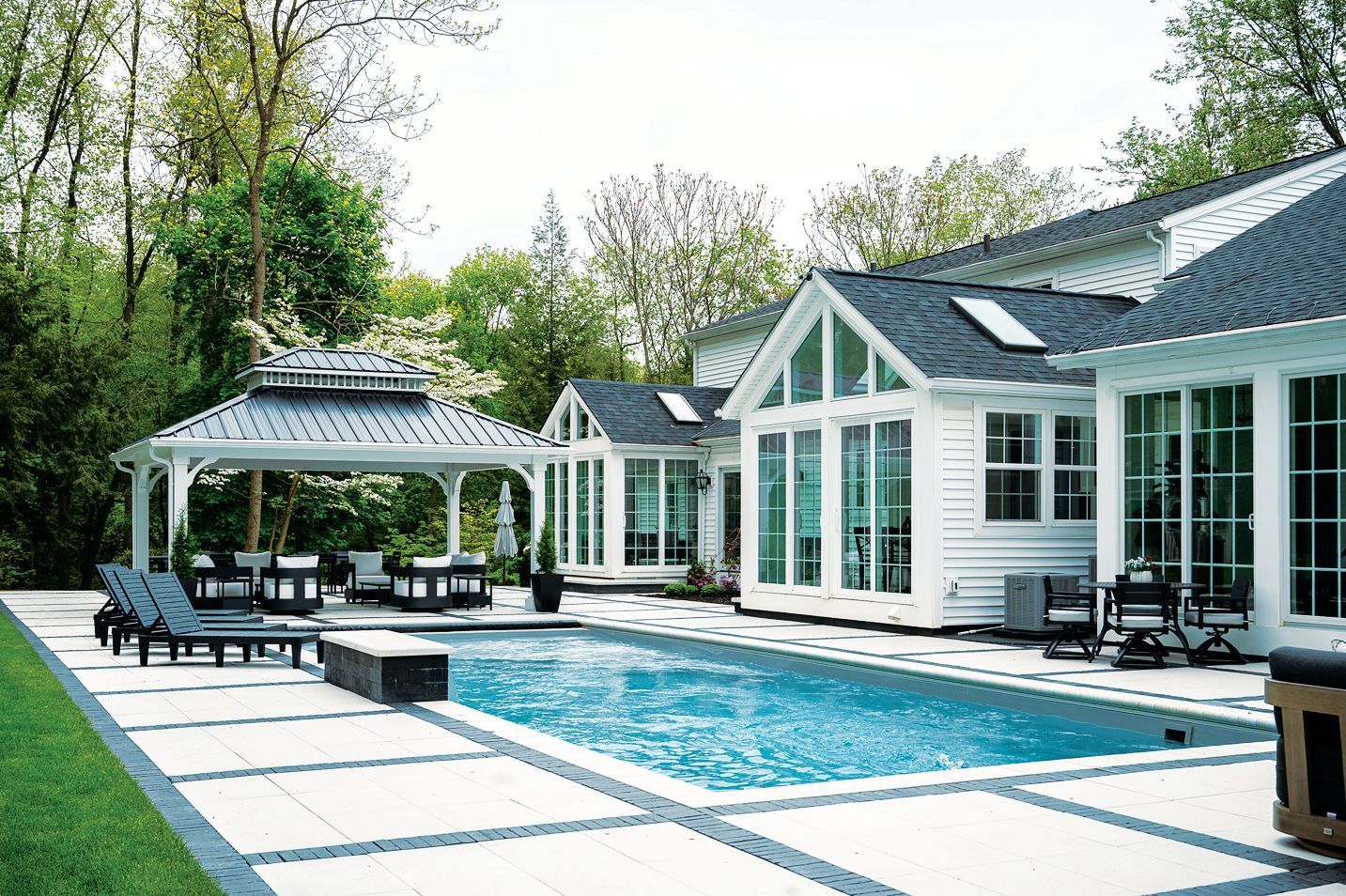 A rectangular swimming pool with a gazebo, patio chairs, and white house with large windows in a lush backyard.