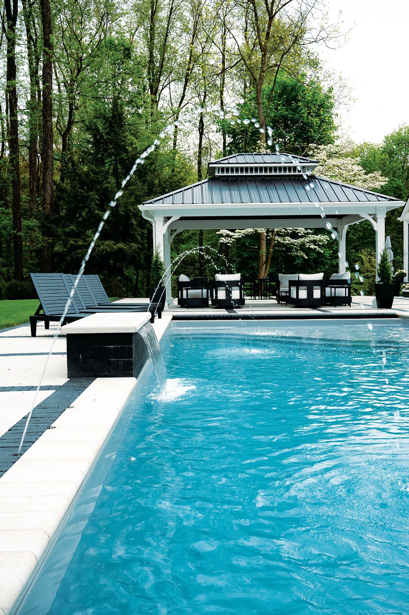 A clear swimming pool with a fountain jet, alongside lounge chairs and a covered pavilion in a leafy, green backyard.