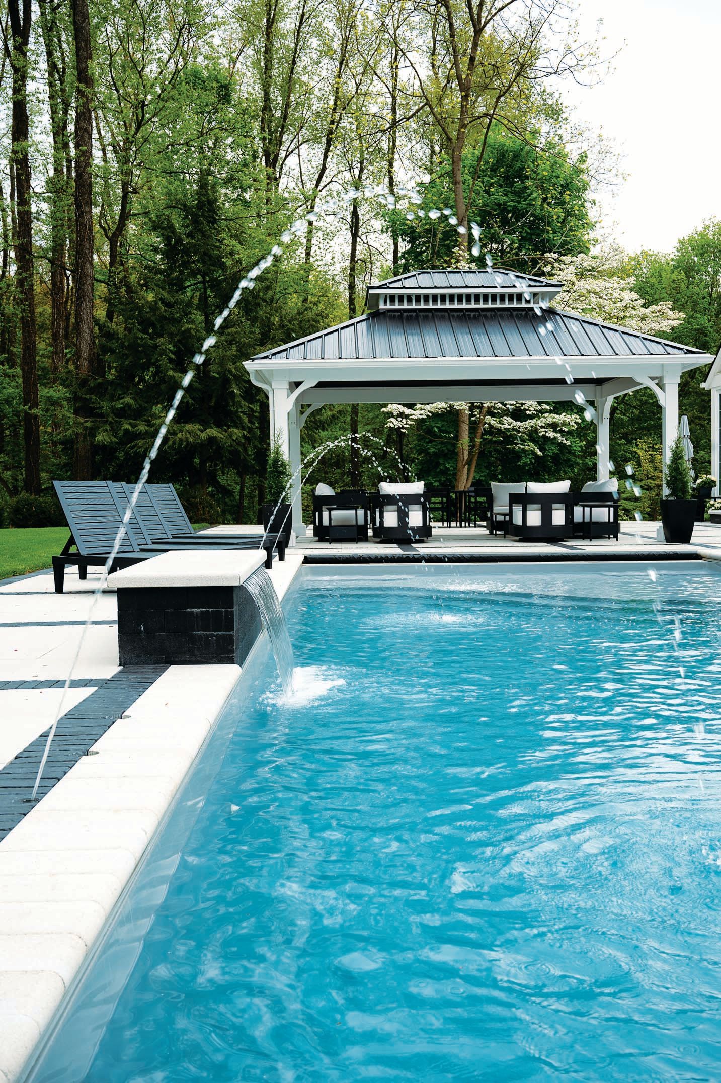 A backyard swimming pool with fountains, lounge chairs, and a white gazebo surrounded by trees.