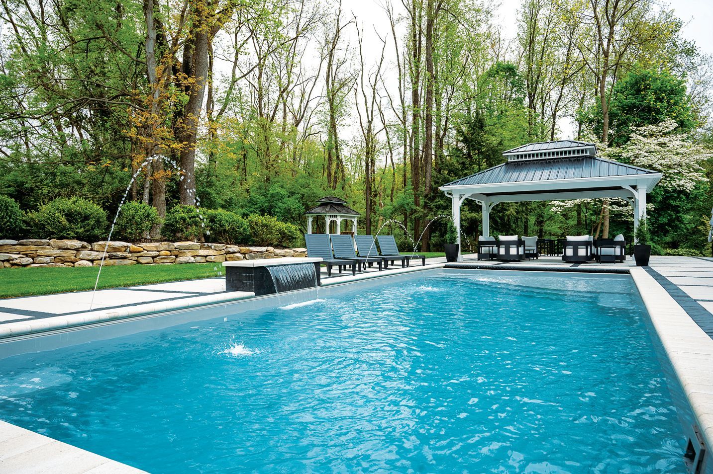 A rectangular swimming pool with bright blue water sits on a patio next to a white gazebo in a wooded backyard.