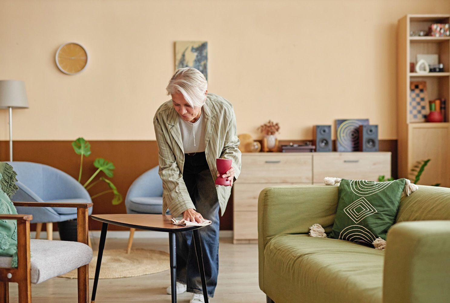 A person wipes a coffee table in a living room with light walls, a green sofa, blue armchairs, and wood furniture.