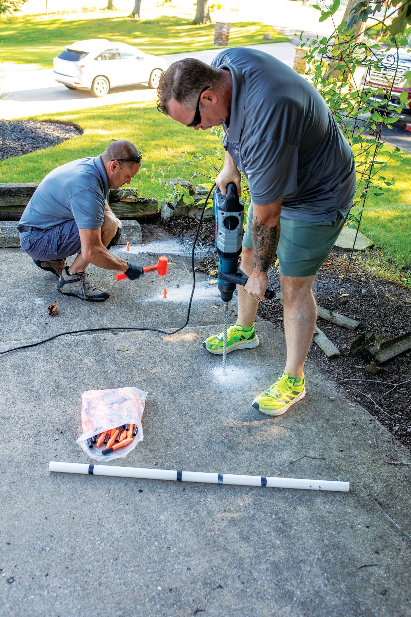 Two people use a rotary hammer and a red mallet to drill and install hardware into a concrete driveway outdoors.