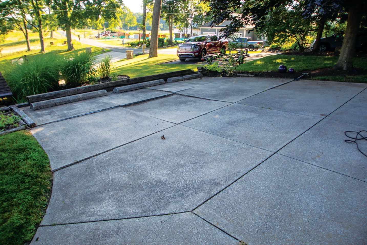 A concrete patio sits in a yard near a driveway, with a red truck visible in the background under large green trees.