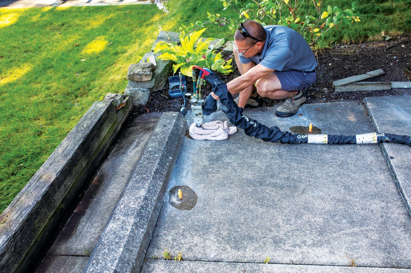 A worker in a blue shirt crouches on a patio, using a drill to inject material into a hole in the concrete.