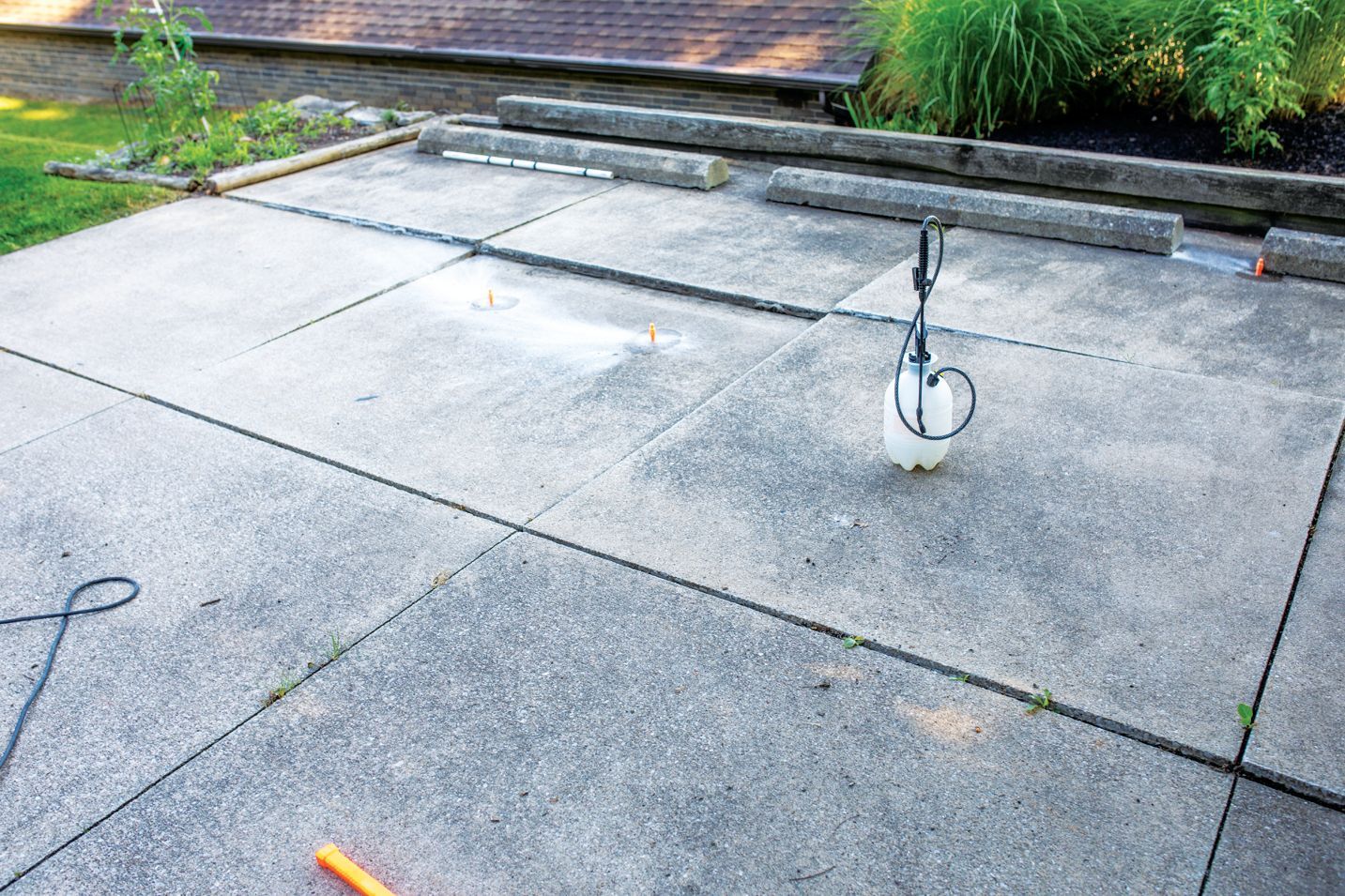 A handheld garden sprayer sits on a gray concrete patio divided into large squares, with wooden landscape timbers nearby.