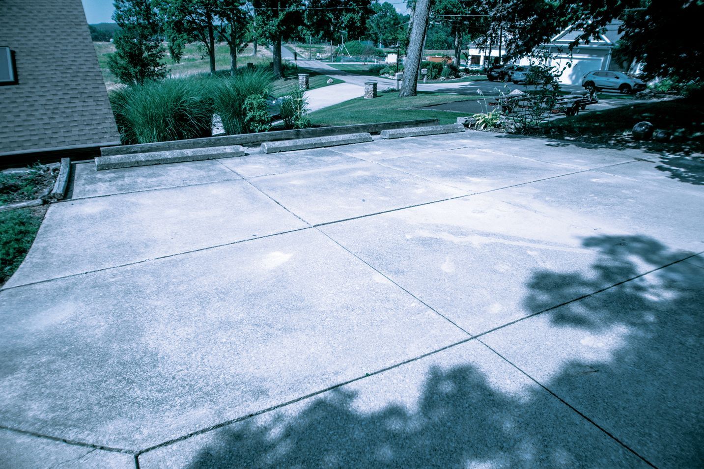 A concrete driveway with a grid pattern of control joints, viewed from the edge of a residential lawn and house.