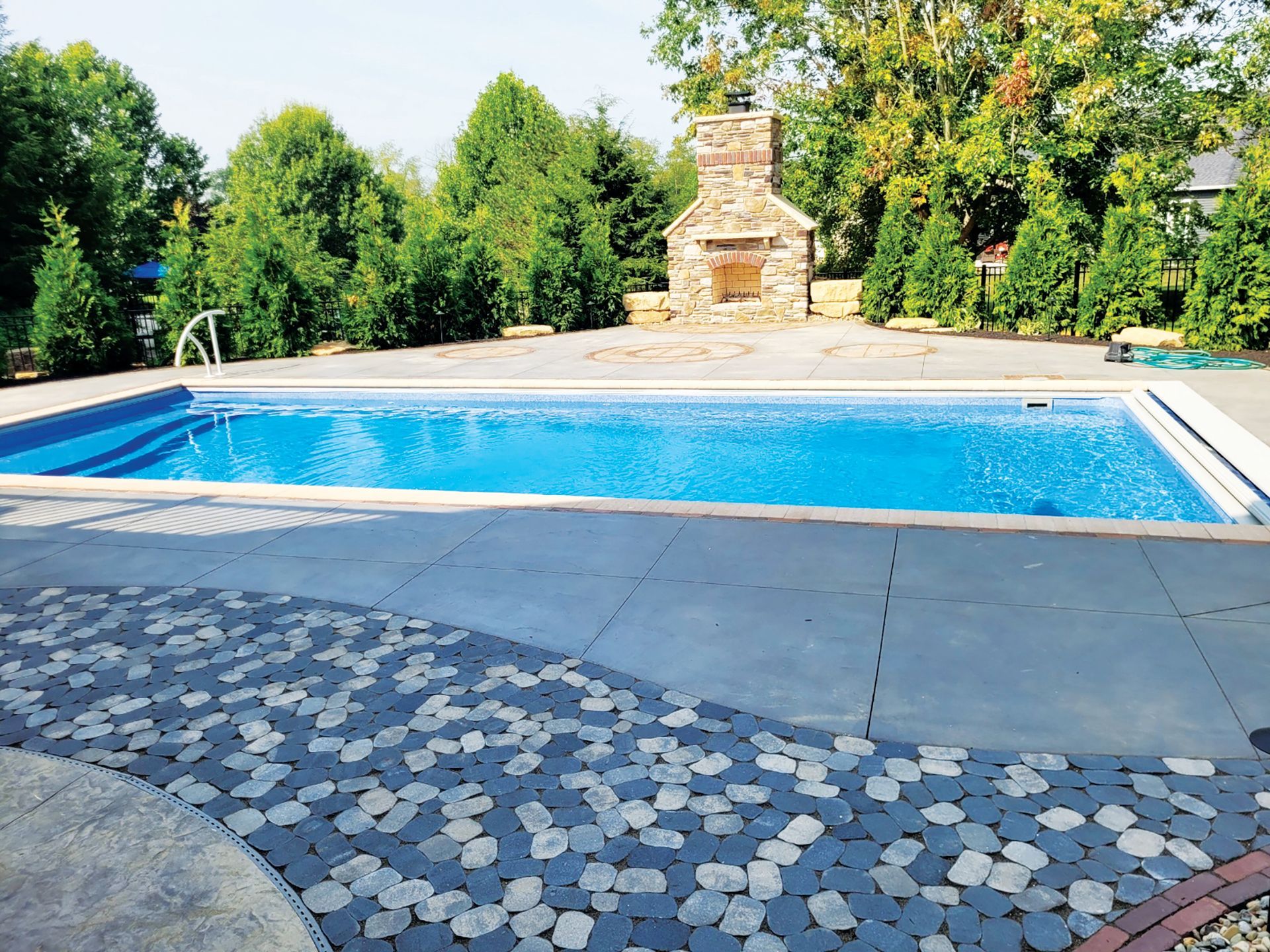 A bright blue rectangular swimming pool with a stone fireplace on the patio, surrounded by trees.