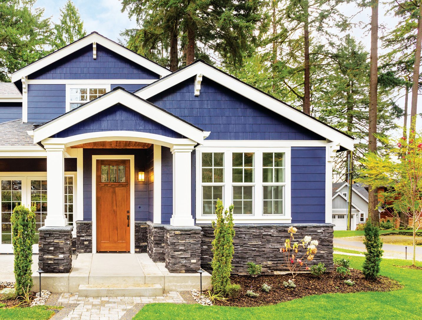 A modern home with dark blue siding, stone accents, a wood front door, and white columns surrounded by trees and greenery.