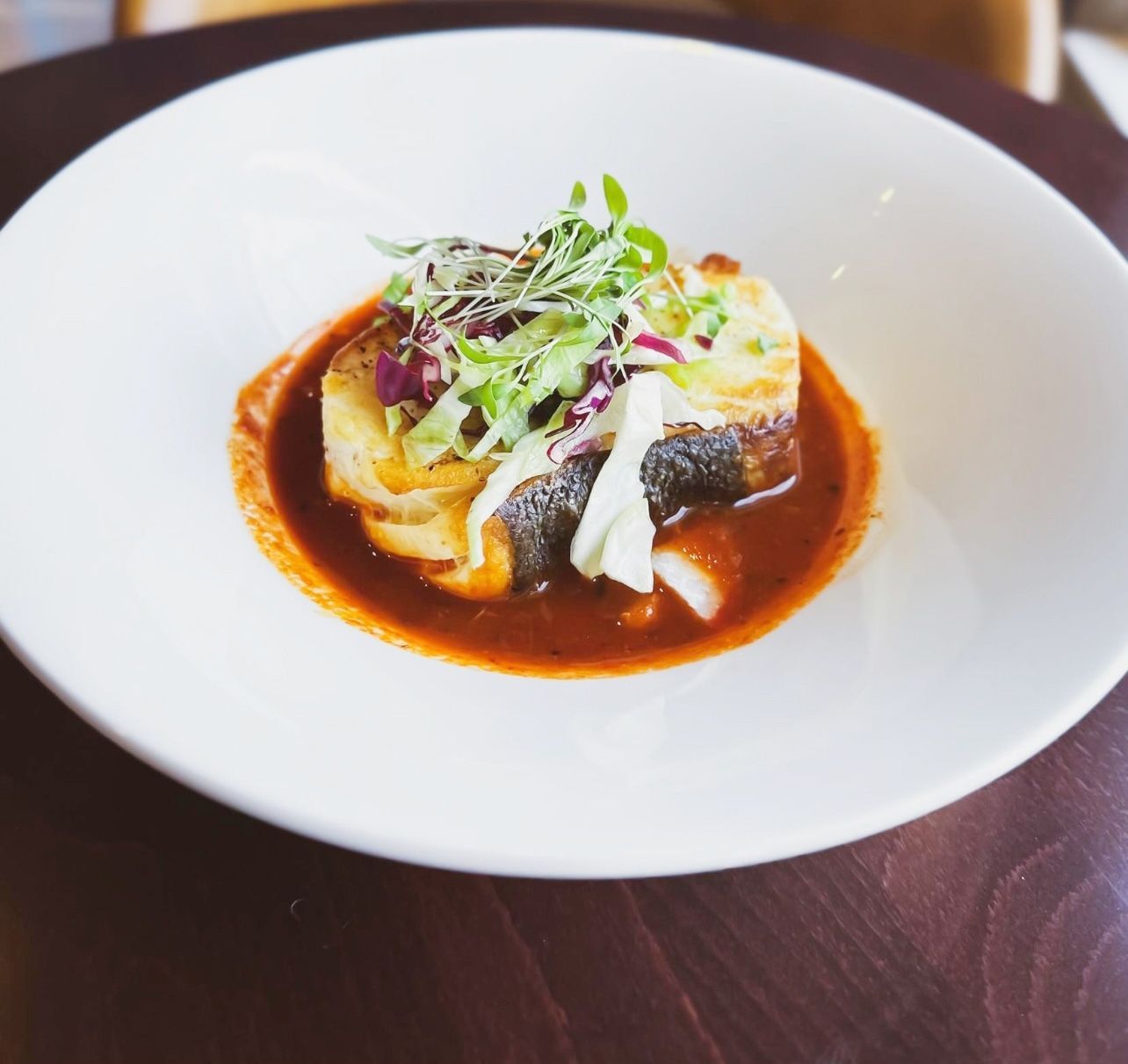 A plated fish dish topped with fresh greens, served in a rich red tomato sauce in a wide white bowl on a wooden table.